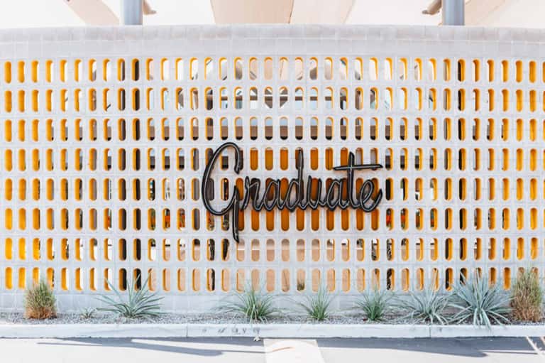 White and yellow perforated wall with 'Graduate' neon sign and desert landscaping at resort entrance