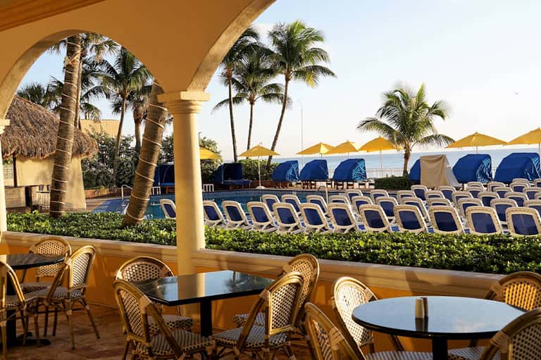 Beachfront dining area with yellow umbrellas, blue lounge chairs, palm trees, and ocean view