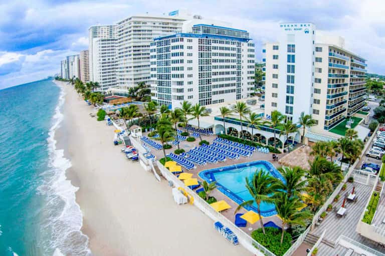 Beachfront resort with white multi-story buildings, blue lounge chairs on sandy beach, ocean waves, and palm trees