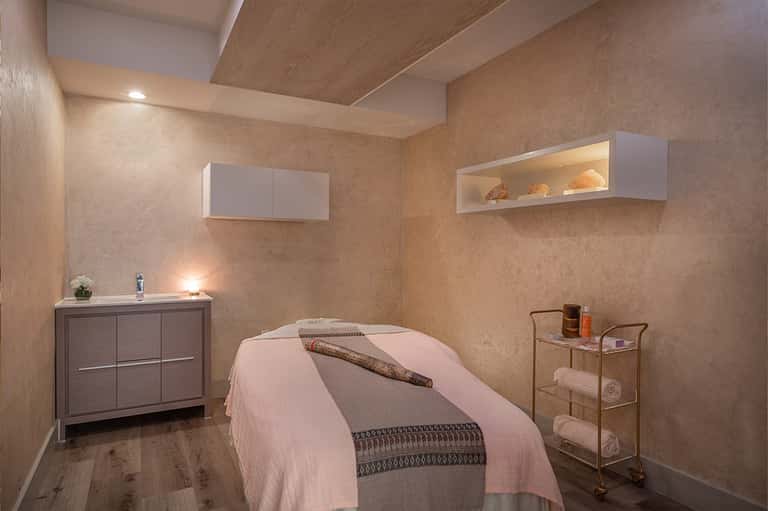 Spa treatment room with warm beige walls, massage bed, wooden storage cabinet, and shelving with folded towels