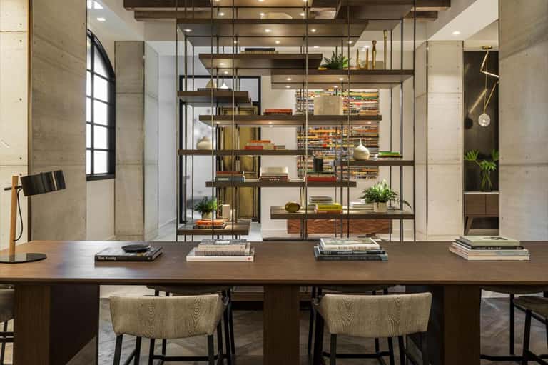 Modern office workspace with wooden desk, woven chairs, and floor-to-ceiling metal shelving displaying books and decorative items