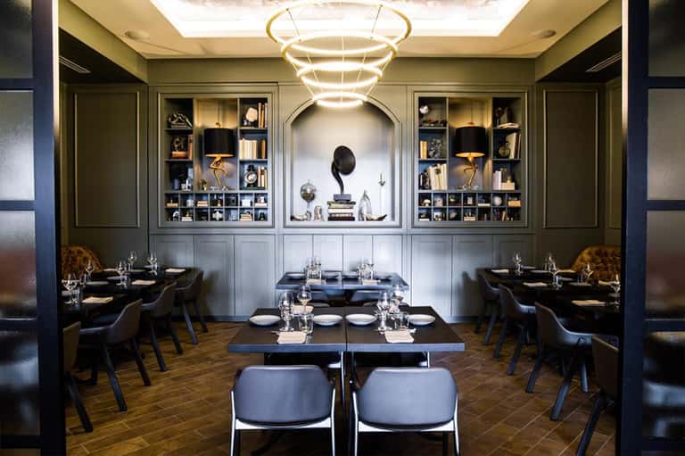 Elegant fine dining restaurant with dark green walls, built-in shelving, circular gold pendant light, and black leather seating