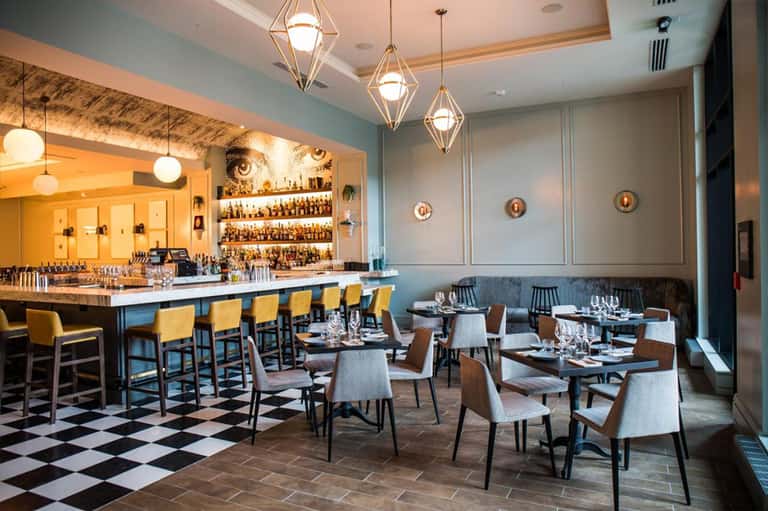 Modern restaurant interior with blue walls, bar seating with yellow chairs, pendant lights, and dining tables