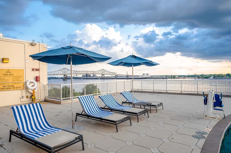 Waterfront deck with blue striped lounge chairs and umbrellas overlooking a bridge and river