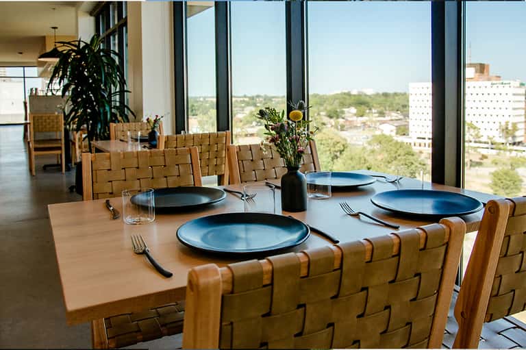 Modern dining room with wooden woven chairs, blue place settings, and panoramic city views through floor-to-ceiling windows
