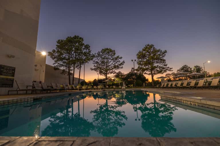 Resort swimming pool at dusk with lounge chairs and tall pine trees reflected in calm water