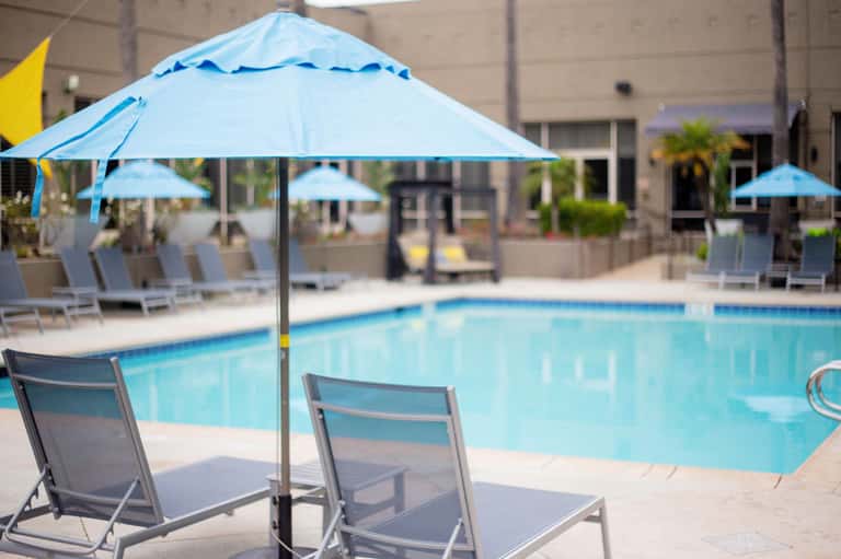 Resort pool with blue umbrellas, lounge chairs, and surrounding buildings on a sunny day