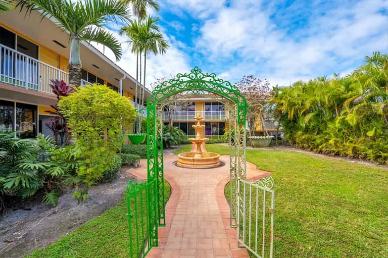 Green ornate garden arbor framing a brick pathway leading to a central fountain with palm trees