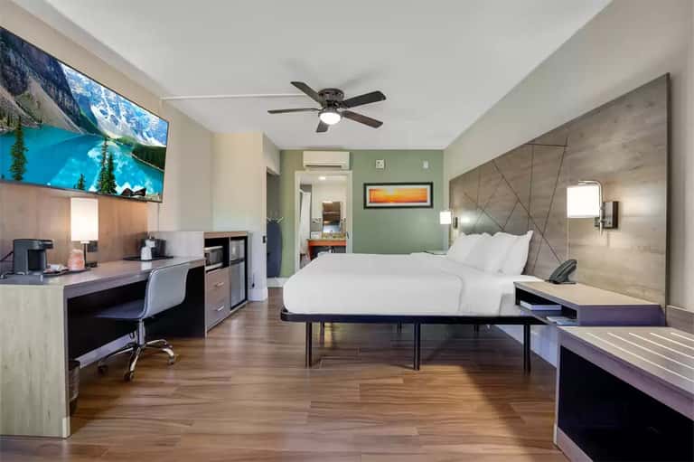 Modern hotel bedroom with king bed, work desk, TV, and green accent wall
