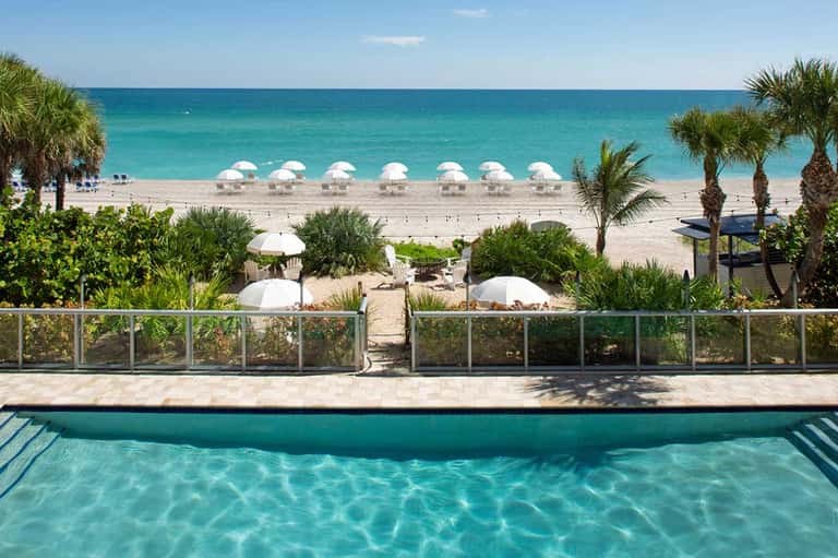 Resort pool with turquoise water overlooking pristine beach with white umbrellas and palm trees