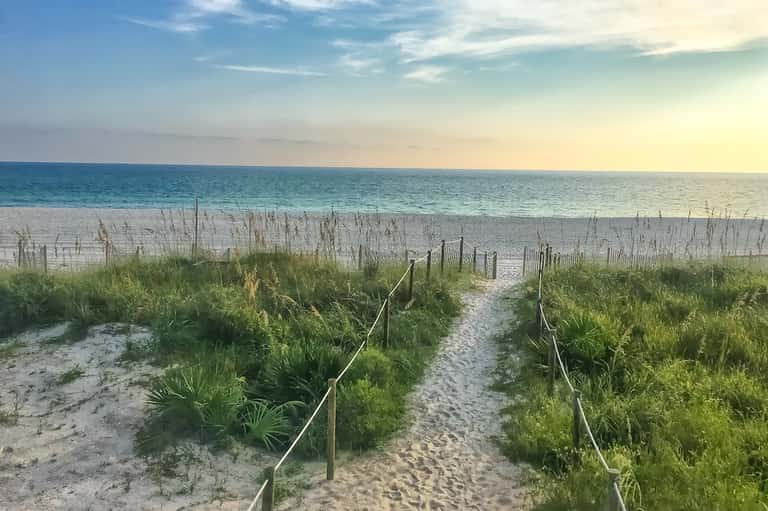 Sandy beach pathway with dune grass, wooden railings, and calm ocean at sunset