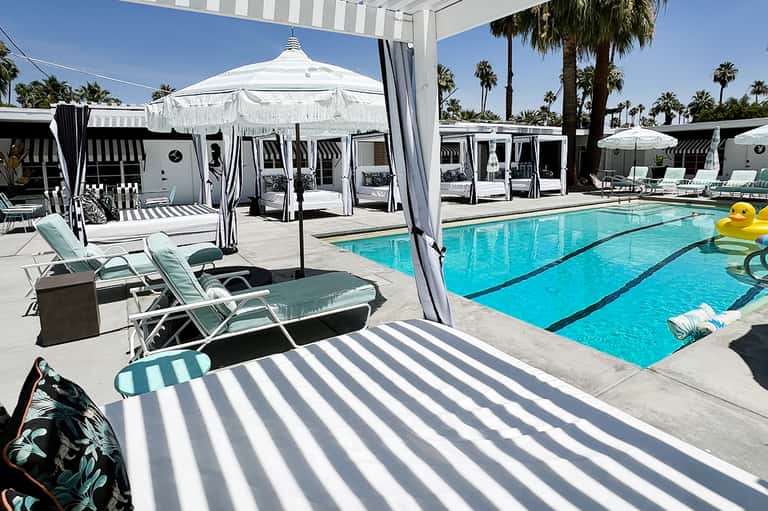 Resort pool with striped lounge chairs, white umbrellas, and palm trees under clear blue sky