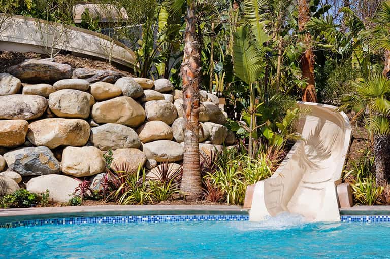Resort pool with waterslide, rocky landscape features, palm trees, and manicured gardens