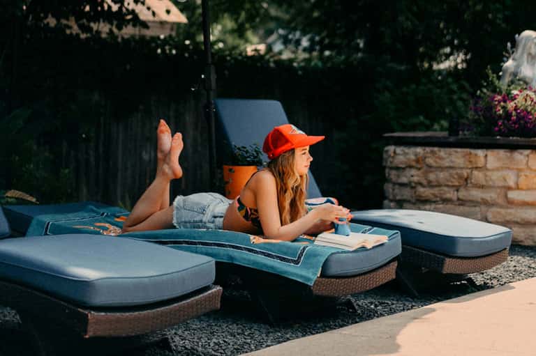 Woman in red cap relaxing on blue lounge chairs by pool in sunny resort setting