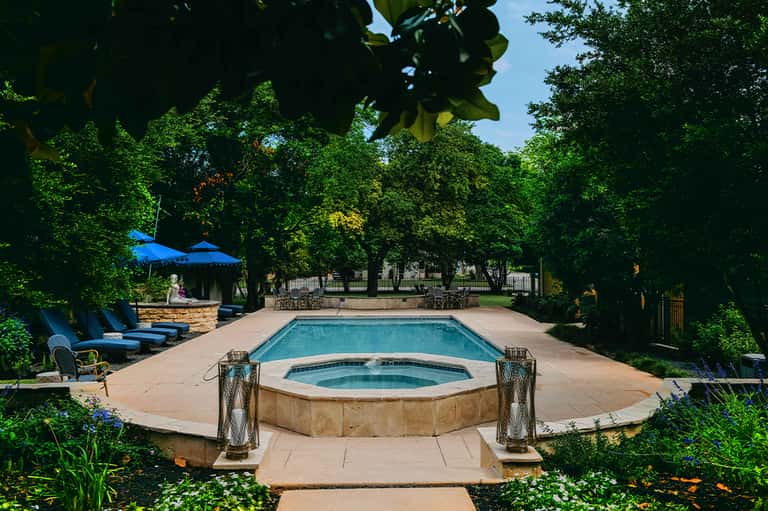 Resort pool with hot tub, loungers, blue umbrellas, and lush trees surrounding patio area