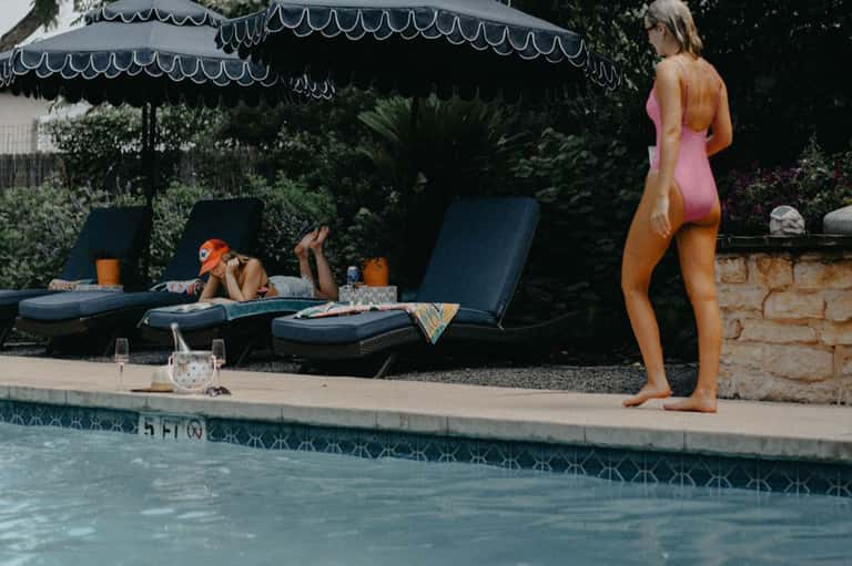 Woman in pink swimsuit standing by resort pool with loungers and umbrellas in background