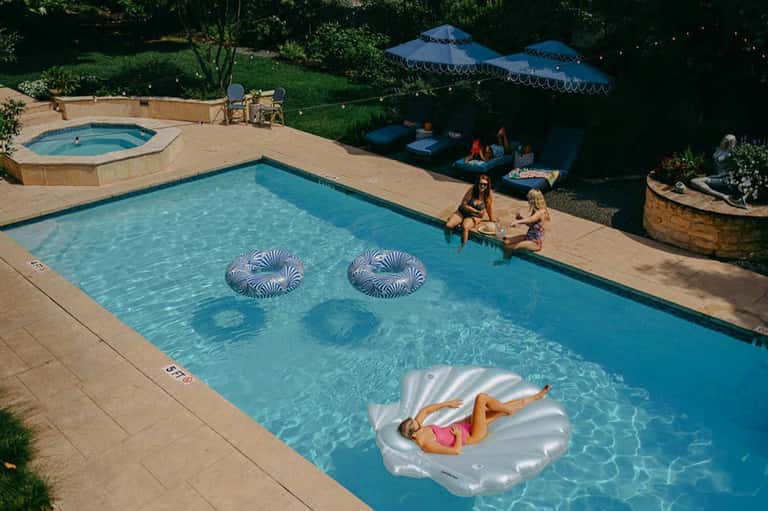 Resort pool with float loungers, spa hot tub, and guests relaxing under blue umbrellas amid lush landscaping