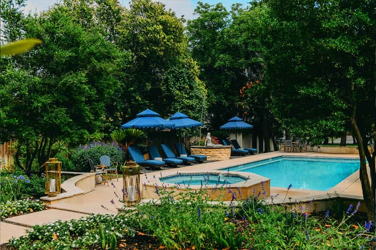 Resort pool area surrounded by blue umbrellas, lounge chairs, and mature trees in natural setting