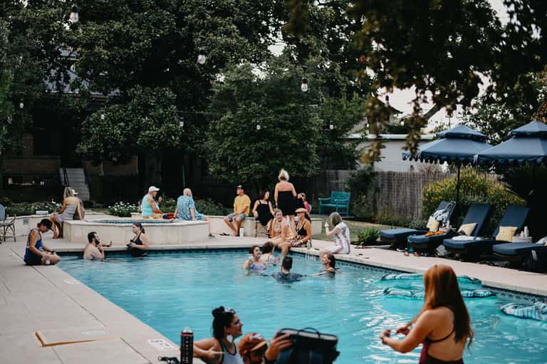 Busy resort pool with guests swimming and relaxing on lounge chairs surrounded by shade umbrellas and mature trees