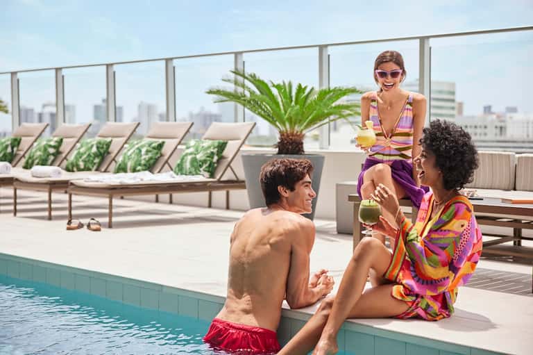 Woman and man enjoying drinks by a rooftop pool with city skyline views, lounge chairs, and palm plants