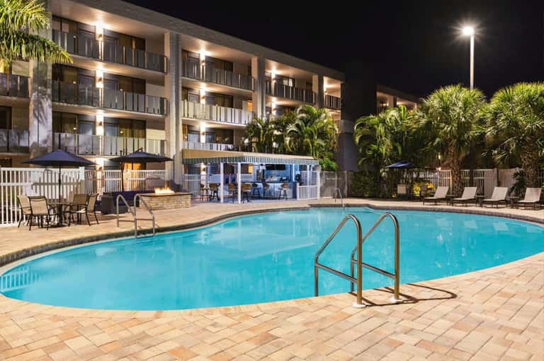 Illuminated resort pool at night with multi-story hotel building, palm trees, and lounge seating area