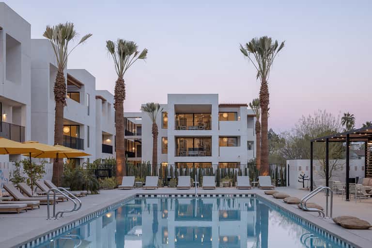 Modern white residential complex with palm trees and illuminated pool at dusk