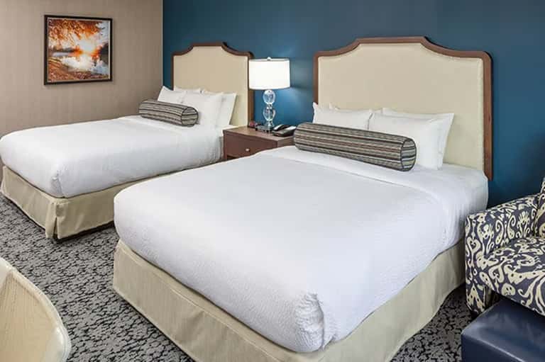 Hotel room with two queen beds, cream upholstered headboards, blue accent wall, and bedside lamp