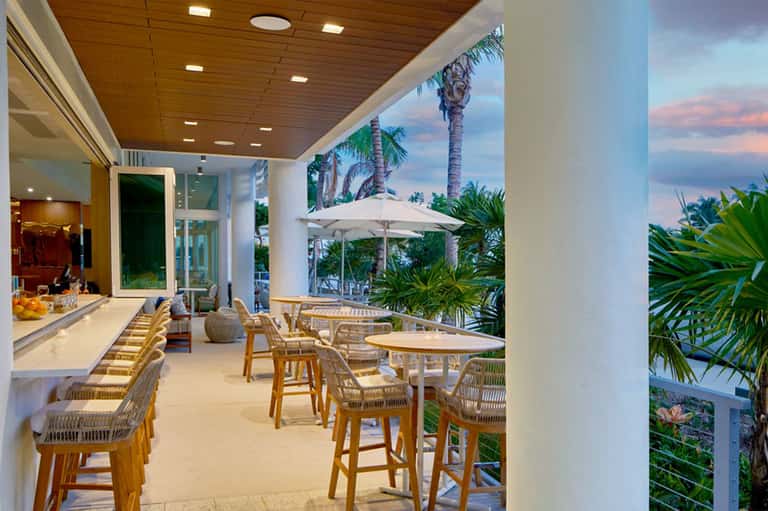 Luxury beachfront terrace with wooden dining chairs, white umbrellas, palm trees, and ocean sunset view