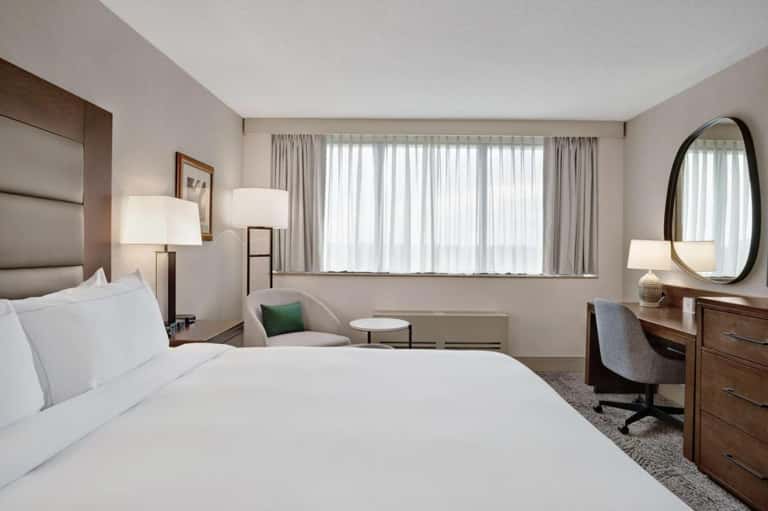 Spacious hotel bedroom with queen bed, work desk, armchair, and large windows with sheer curtains