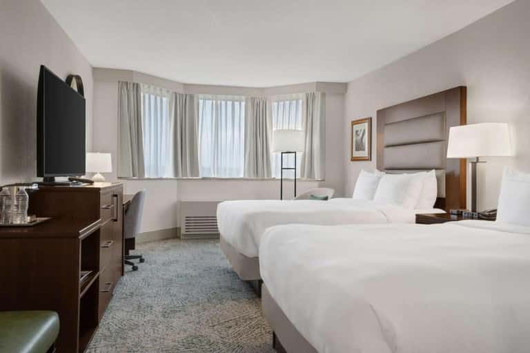 Hotel bedroom with two queen beds, wood furniture, and bay windows with curtains