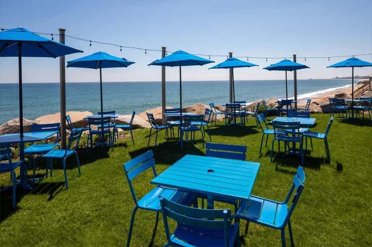Beachfront dining area with blue umbrellas and tables on grass overlooking ocean and sandy shore