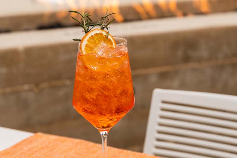 Orange cocktail with rosemary garnish and orange slice on poolside table