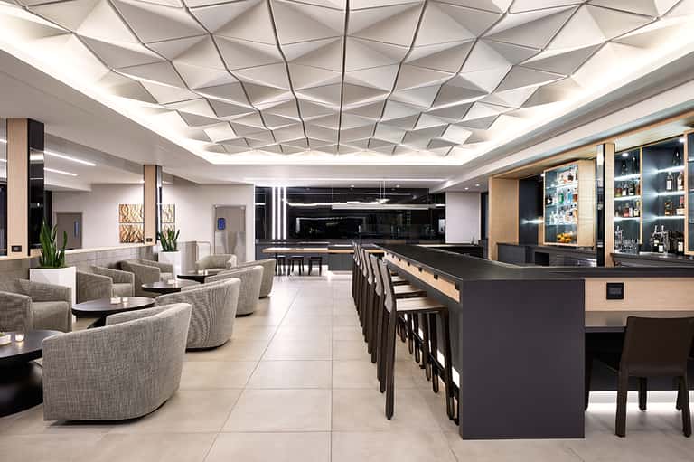 Modern hotel bar and lounge with geometric ceiling design, beige seating, and black bar counter