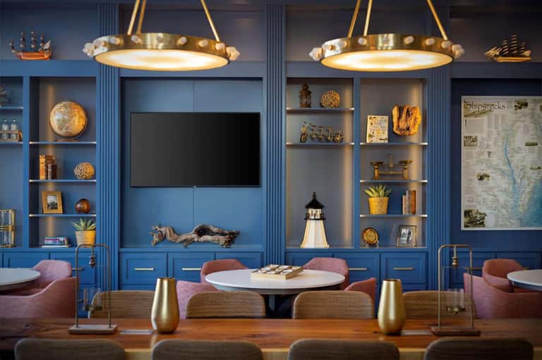 Upscale lounge with blue cabinetry, brass pendant lights, round tables with pink chairs, and nautical-themed shelving