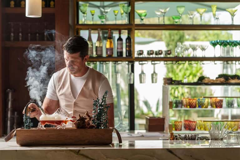 Man performing tea ceremony at luxury bar with botanical garden views and backlit glassware shelving