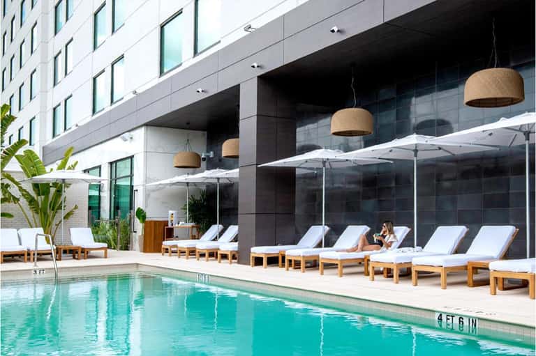 Resort pool with turquoise water, white umbrellas, wooden loungers, and glass building backdrop