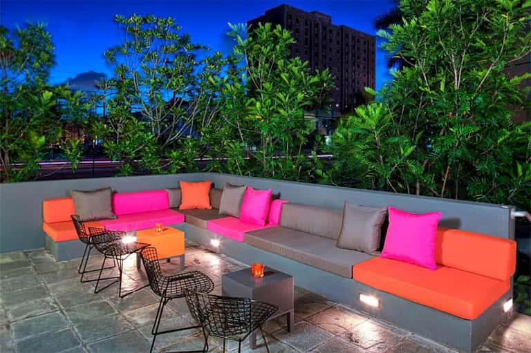 Rooftop lounge at dusk with colorful cushions, potted plants, and city skyline