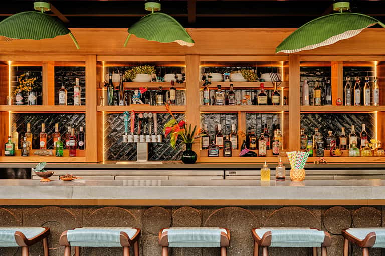 Modern bar with wooden shelving displaying spirits, green leaf-shaped lamps, and striped stools