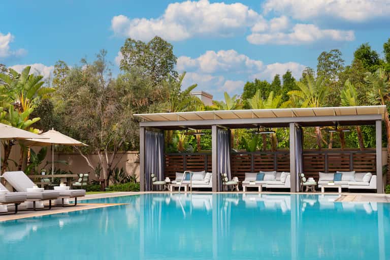 Resort pool with white cabanas, wooden pergolas with curtains, lounge chairs, and lush tropical landscaping