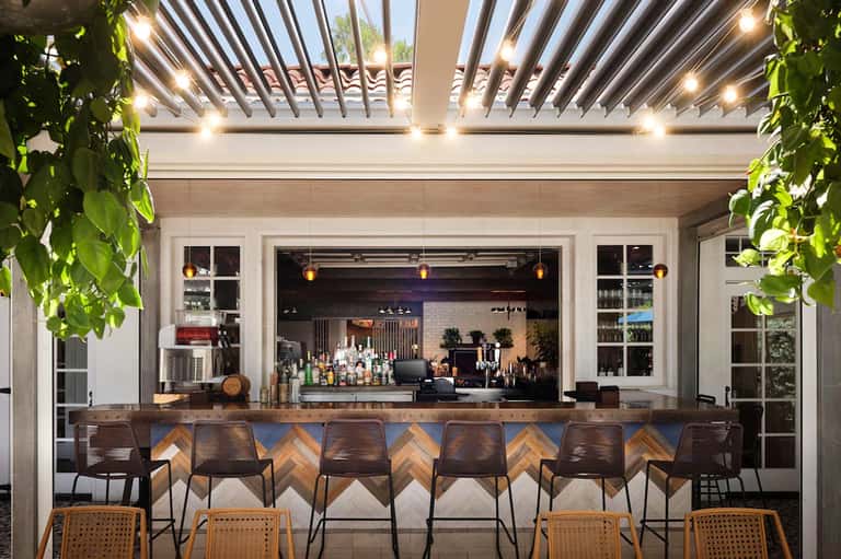 Modern outdoor bar area with wooden pergola ceiling, pendant lights, bar seating, and shelving displaying bottles and glassware