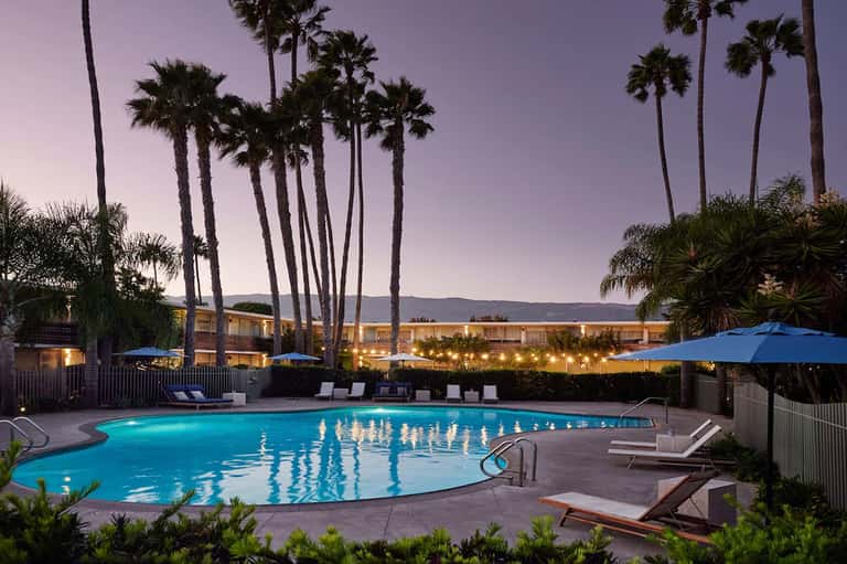 Lit swimming pool at dusk surrounded by palm trees and blue umbrellas with resort buildings in background