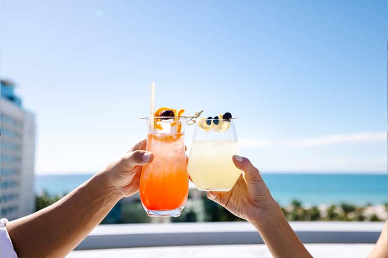 Two tropical cocktails toasting against ocean and sky backdrop with decorative fruit garnishes