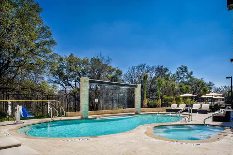 Resort swimming pool with hot tub, lounge chairs, umbrellas, and tree-lined landscape under clear sky