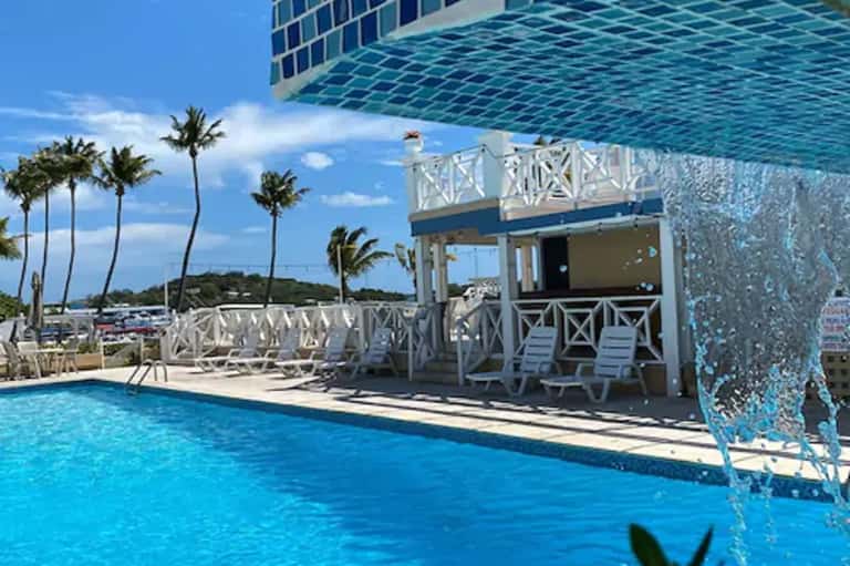 Resort pool with turquoise water, white railings, palm trees, and tropical beach bar with ocean views