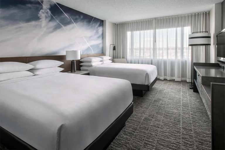 Modern hotel room with two queen beds, floor-to-ceiling windows, and cloud artwork on accent wall