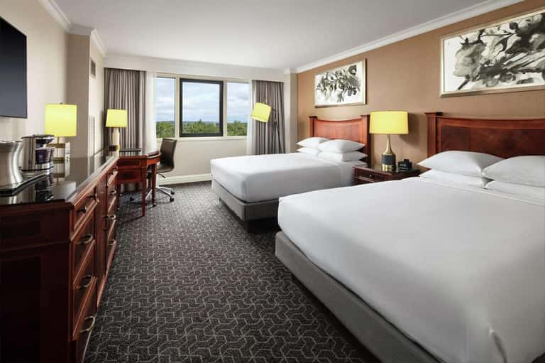 Upscale hotel room with two queen beds, wooden furniture, and panoramic window view of trees