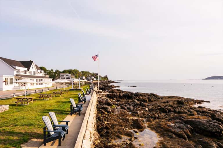 Coastal waterfront with Adirondack chairs on lawn, white buildings, rocky shoreline, and ocean view