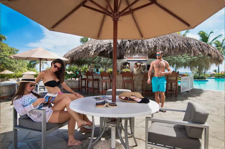 Beachfront bar area with thatched roof, guests relaxing under umbrellas, turquoise water and palm trees visible