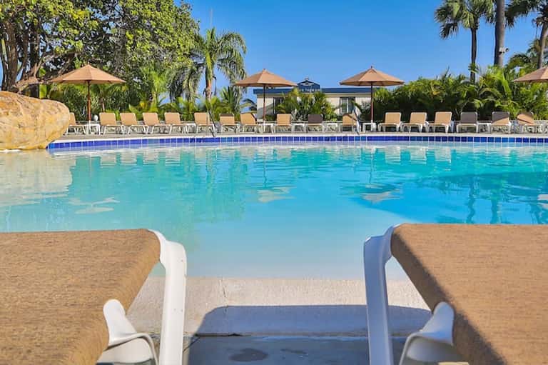 Resort swimming pool with blue water, lounge chairs, umbrellas, and palm trees under clear sky