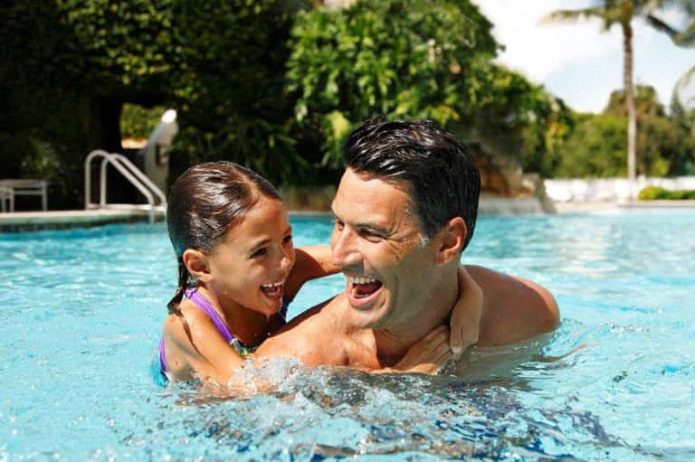 Father and daughter swimming together in bright blue resort pool with tropical landscaping